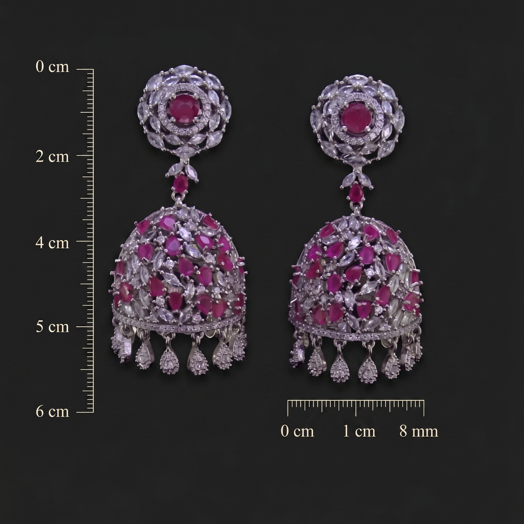 Pair of ornate earrings with gemstones on a dark background with measurement scale.